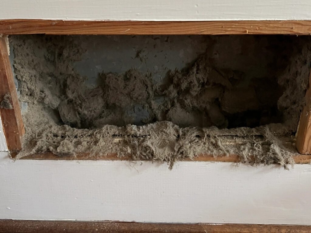 Thick dust and debris buildup around the vent opening and inside the duct, creating a messy and restricted airflow area.
